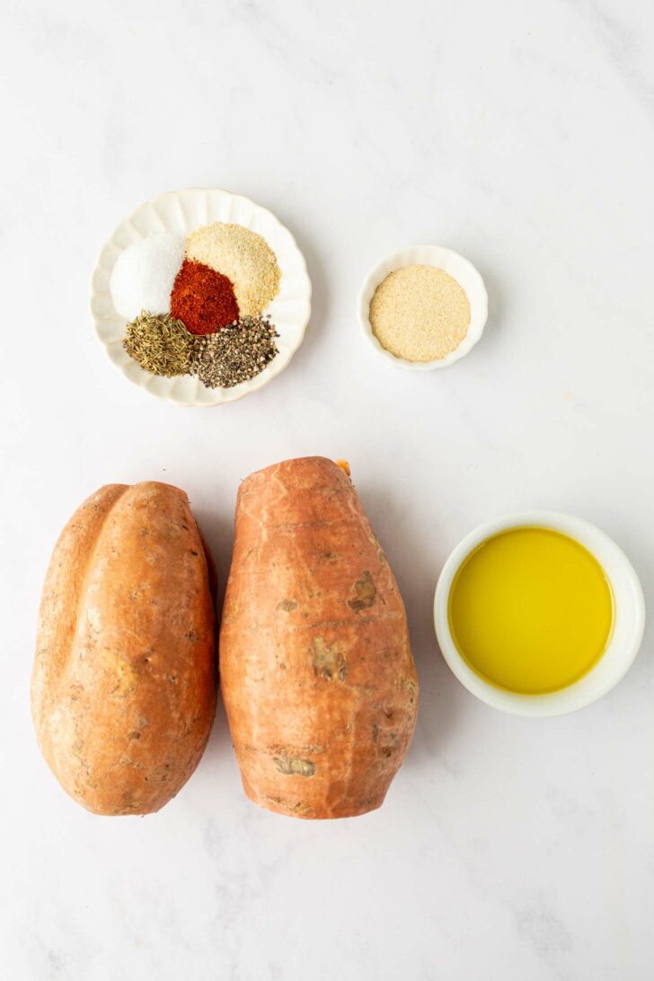 Two whole sweet potatoes, a small bowl of olive oil, a small dish with seasonings (salt, pepper, paprika, garlic powder, dried thyme), and a small bowl of breadcrumbs are arranged on a white surface.
