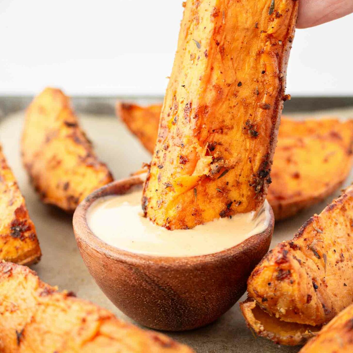 A hand dips a seasoned sweet potato wedge into a small wooden bowl of creamy dipping sauce, with more sweet potato wedges arranged around the bowl.