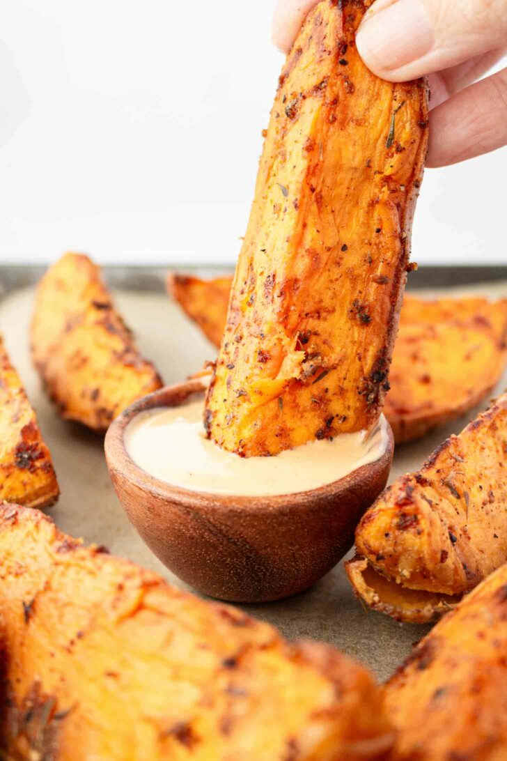 A hand dips a seasoned sweet potato wedge into a small wooden bowl of creamy dipping sauce, with more sweet potato wedges arranged around the bowl.