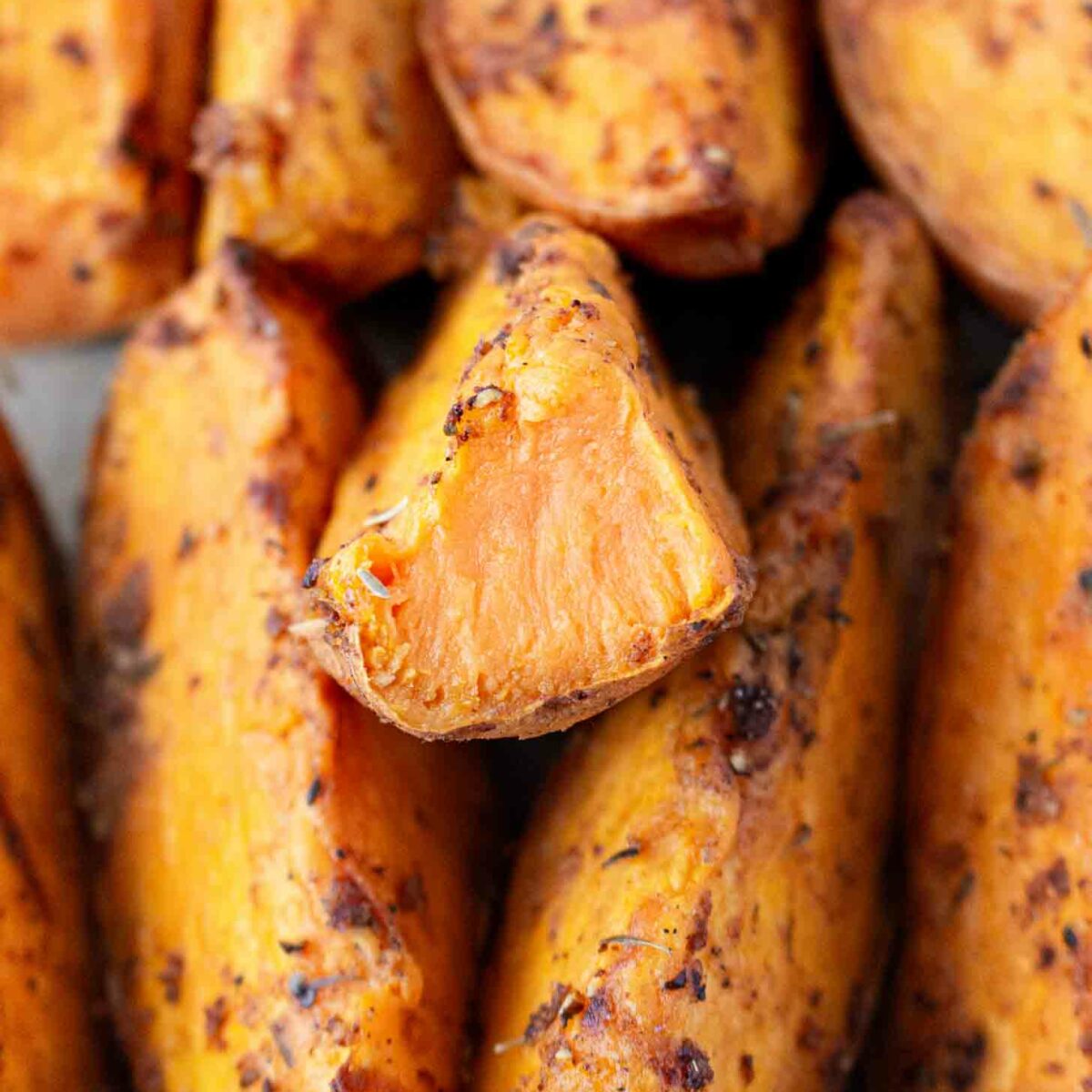 A close-up of roasted sweet potato wedges, seasoned with spices, showing a crispy exterior and soft, orange interior. One wedge is cut to reveal its tender inside.