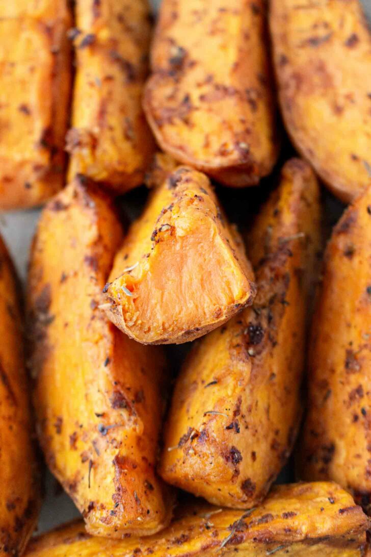 A close-up of roasted sweet potato wedges, seasoned with spices, showing a crispy exterior and soft, orange interior. One wedge is cut to reveal its tender inside.