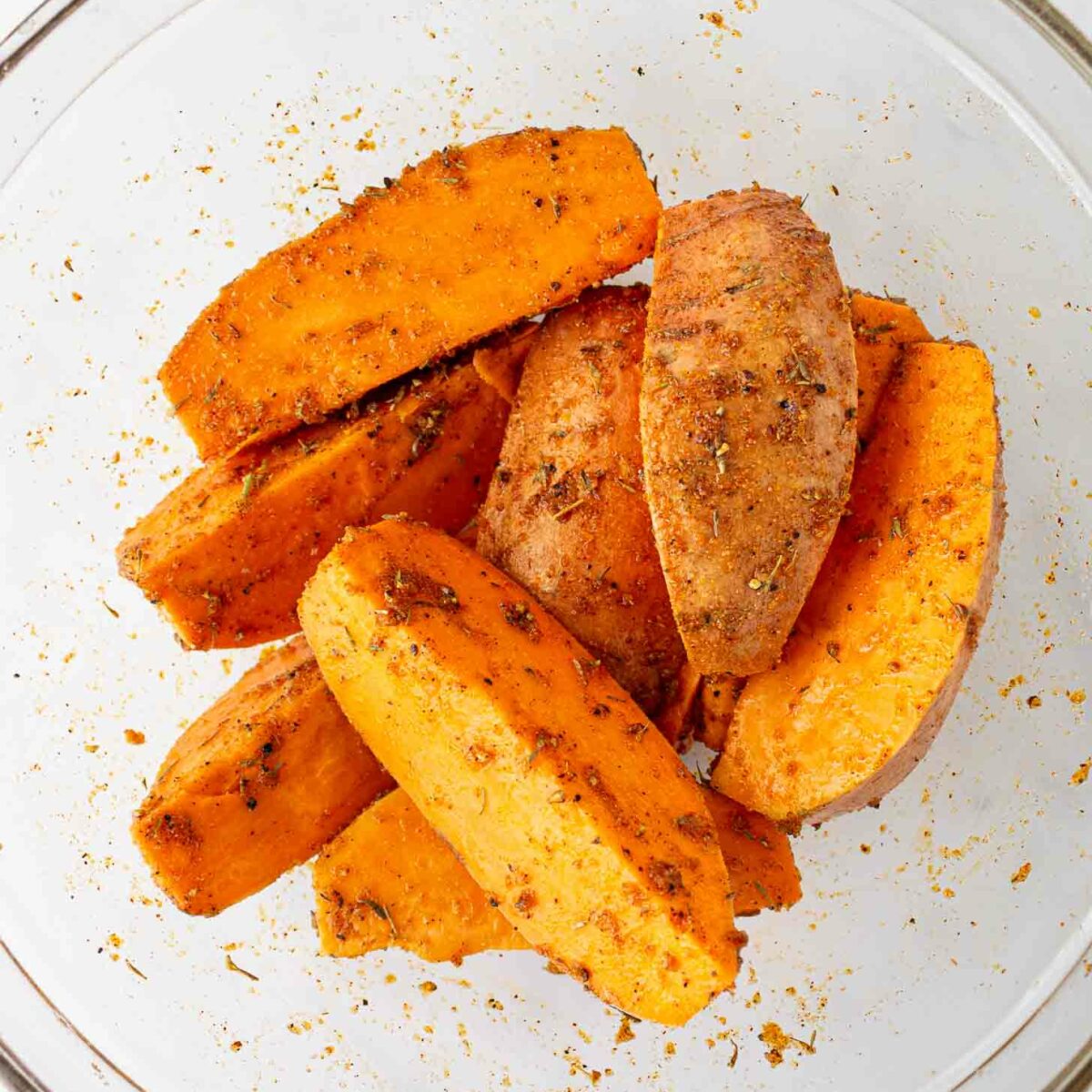 Sweet potato wedges coated with spices and herbs in a clear glass bowl, ready to be baked or cooked.