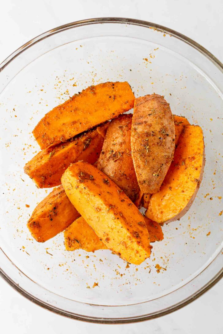 Sweet potato wedges coated with spices and herbs in a clear glass bowl, ready to be baked or cooked.