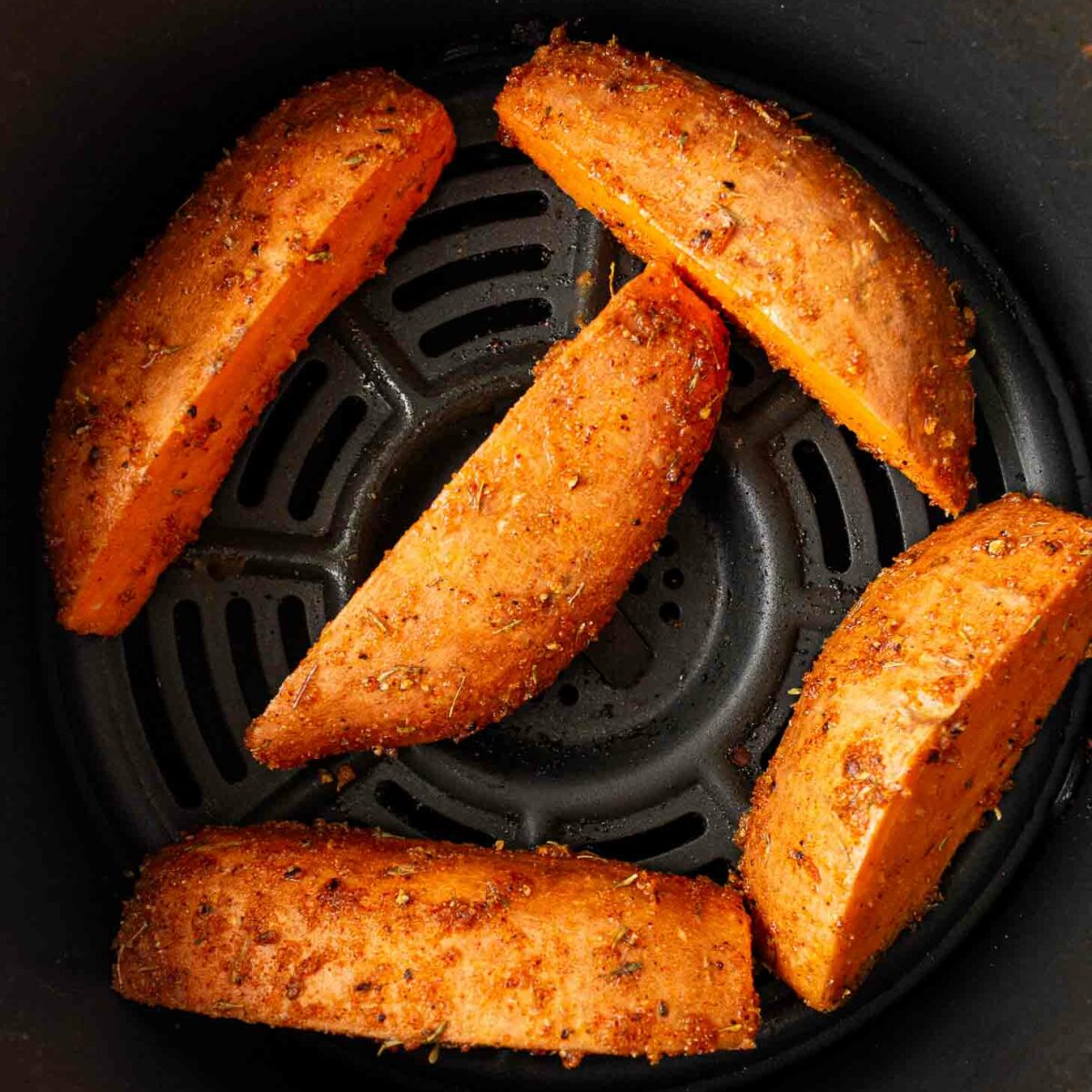Five seasoned sweet potato wedges arranged in a single layer inside an air fryer basket, ready to be cooked.
