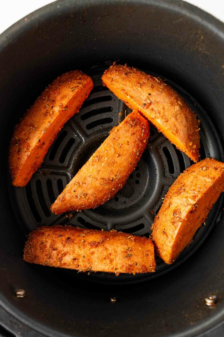 Five seasoned sweet potato wedges arranged in a single layer inside an air fryer basket, ready to be cooked.