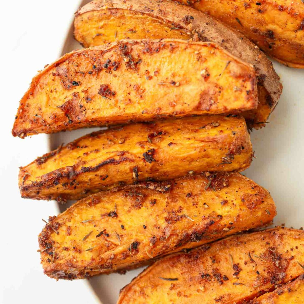 Close-up of several seasoned, roasted sweet potato wedges arranged on a white plate. The wedges have a crispy, golden-brown exterior with visible spices and herbs.