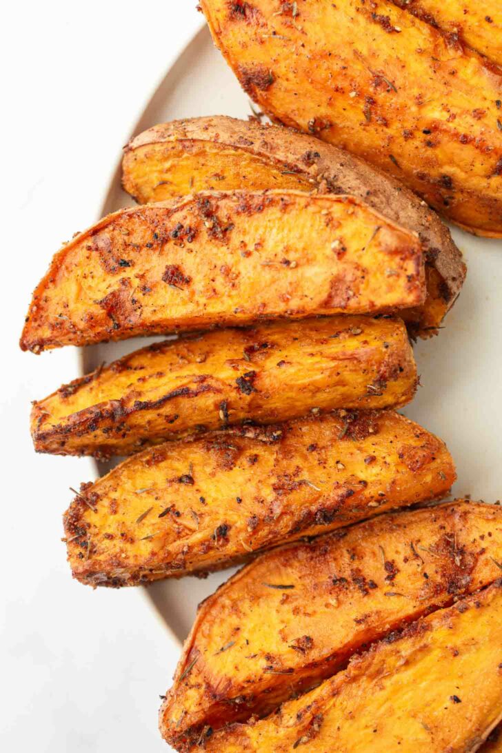 Close-up of several seasoned, roasted sweet potato wedges arranged on a white plate. The wedges have a crispy, golden-brown exterior with visible spices and herbs.