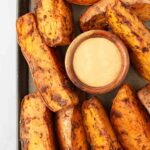 Thick, seasoned sweet potato wedges arranged around a small wooden bowl of creamy dipping sauce on a baking tray.