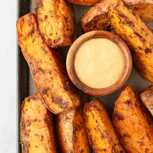 Thick, seasoned sweet potato wedges arranged around a small wooden bowl of creamy dipping sauce on a baking tray.