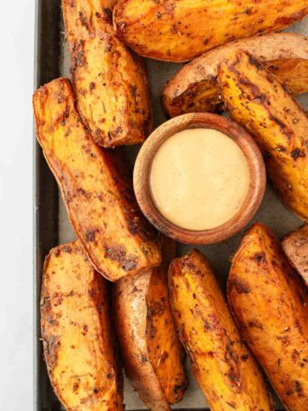 Thick, seasoned sweet potato wedges arranged around a small wooden bowl of creamy dipping sauce on a baking tray.