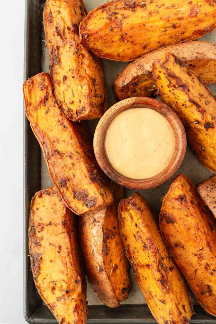 Thick, seasoned sweet potato wedges arranged around a small wooden bowl of creamy dipping sauce on a baking tray.