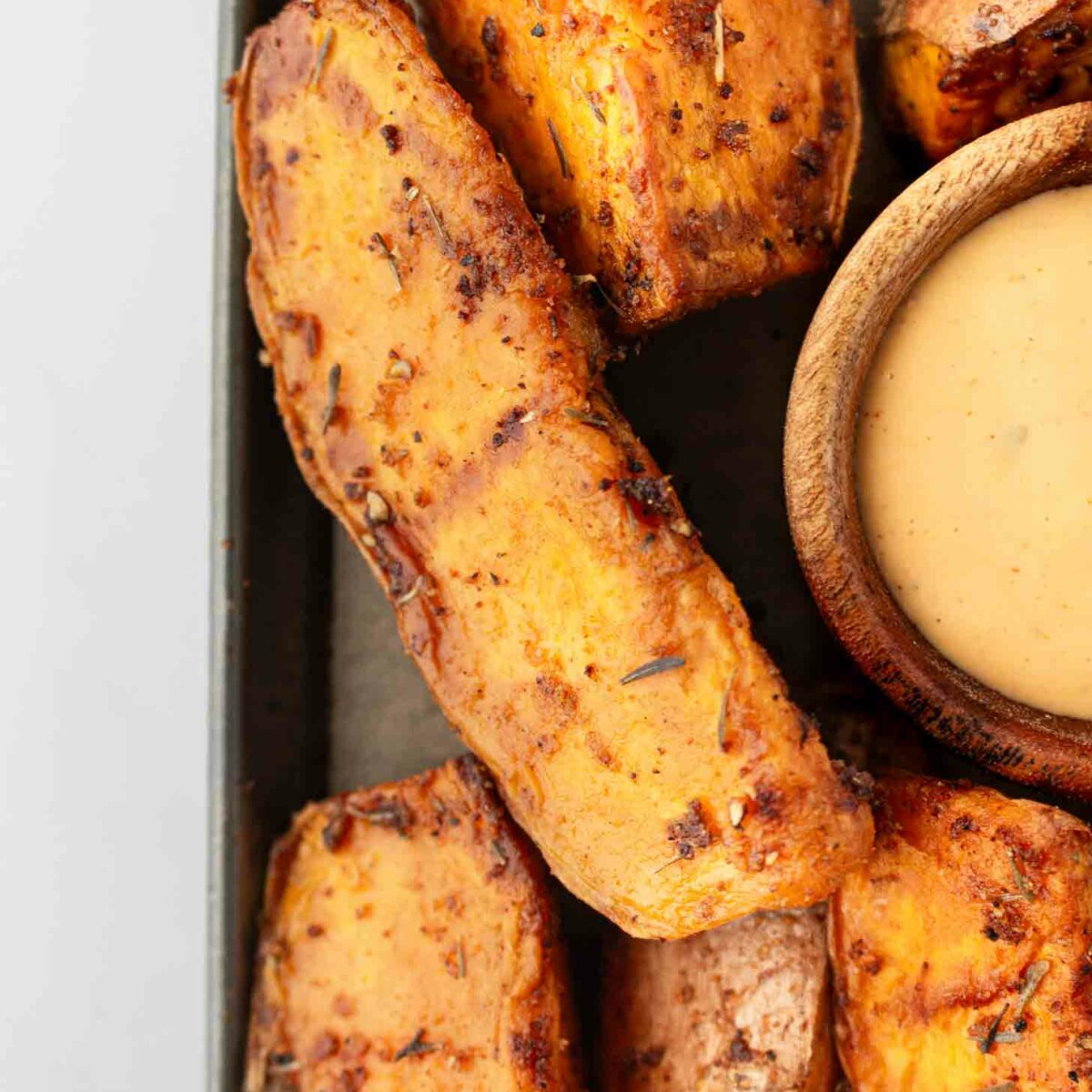 Close-up of seasoned, roasted sweet potato wedges arranged next to a small wooden bowl filled with creamy dipping sauce on a tray.