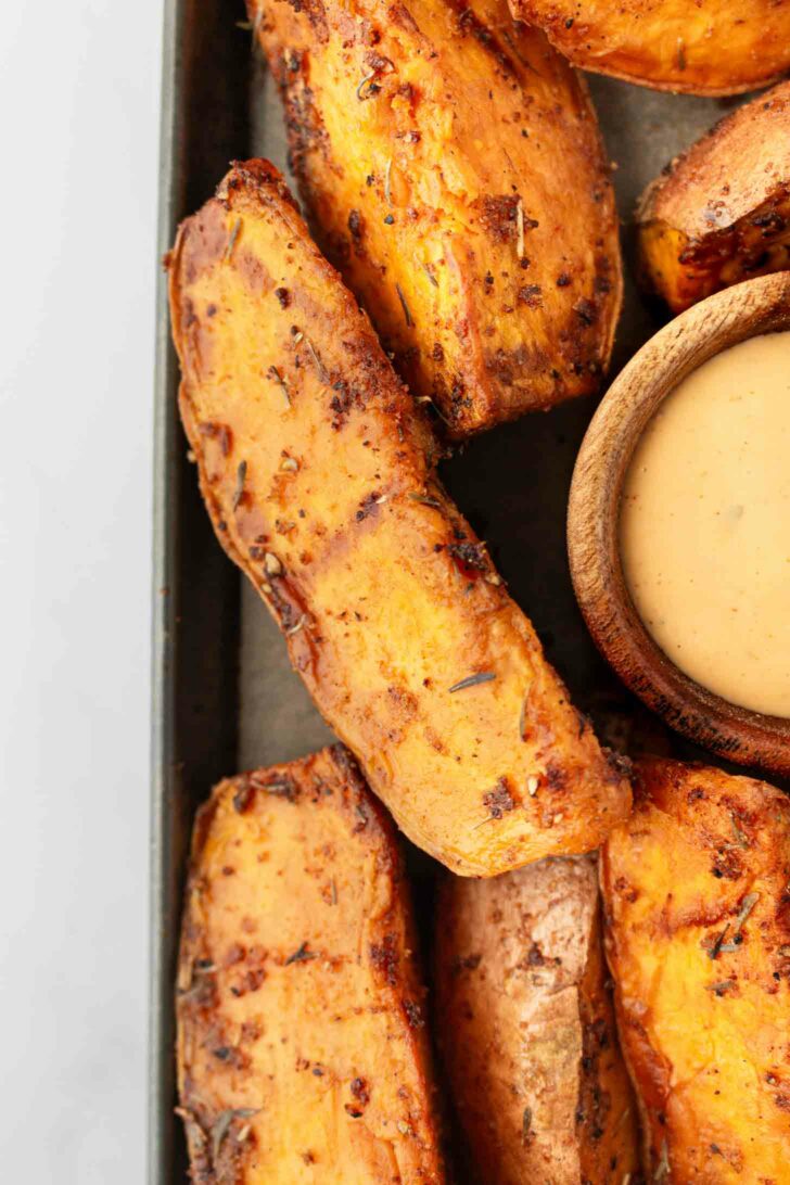 Close-up of seasoned, roasted sweet potato wedges arranged next to a small wooden bowl filled with creamy dipping sauce on a tray.