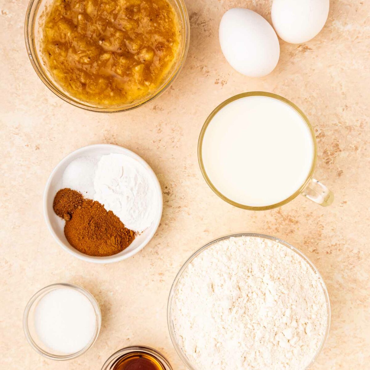 Top-down view of baking ingredients on a beige surface: two eggs, a bowl of mashed bananas, a cup of milk, a bowl of flour, a bowl with spices and baking powder, a small bowl of sugar, and a small dish of vanilla extract.