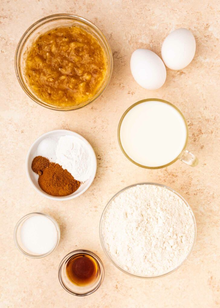 Top-down view of baking ingredients on a beige surface: two eggs, a bowl of mashed bananas, a cup of milk, a bowl of flour, a bowl with spices and baking powder, a small bowl of sugar, and a small dish of vanilla extract.