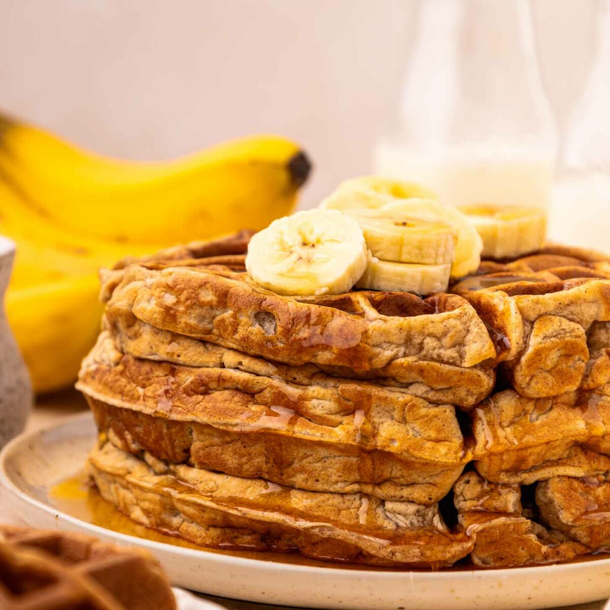 A stack of waffles topped with banana slices and drizzled with syrup sits on a plate. In the background, there are bananas and glass bottles with milk.
