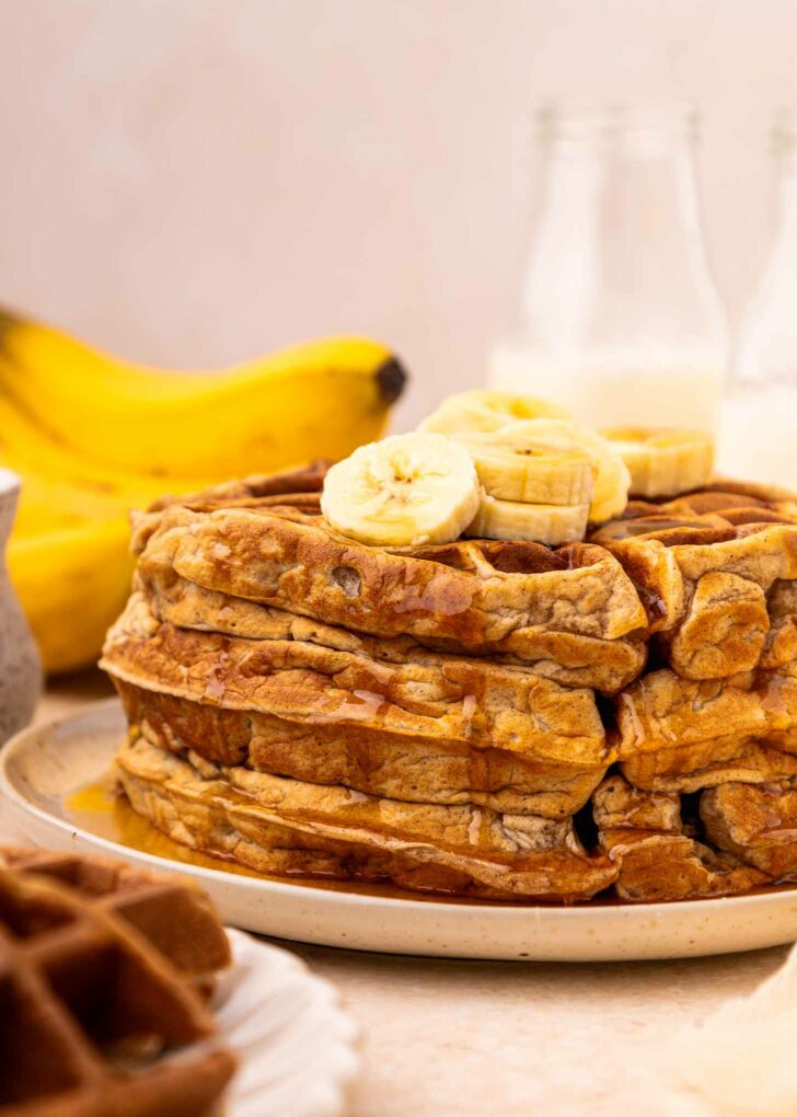 A stack of waffles topped with banana slices and drizzled with syrup sits on a plate. In the background, there are bananas and glass bottles with milk.