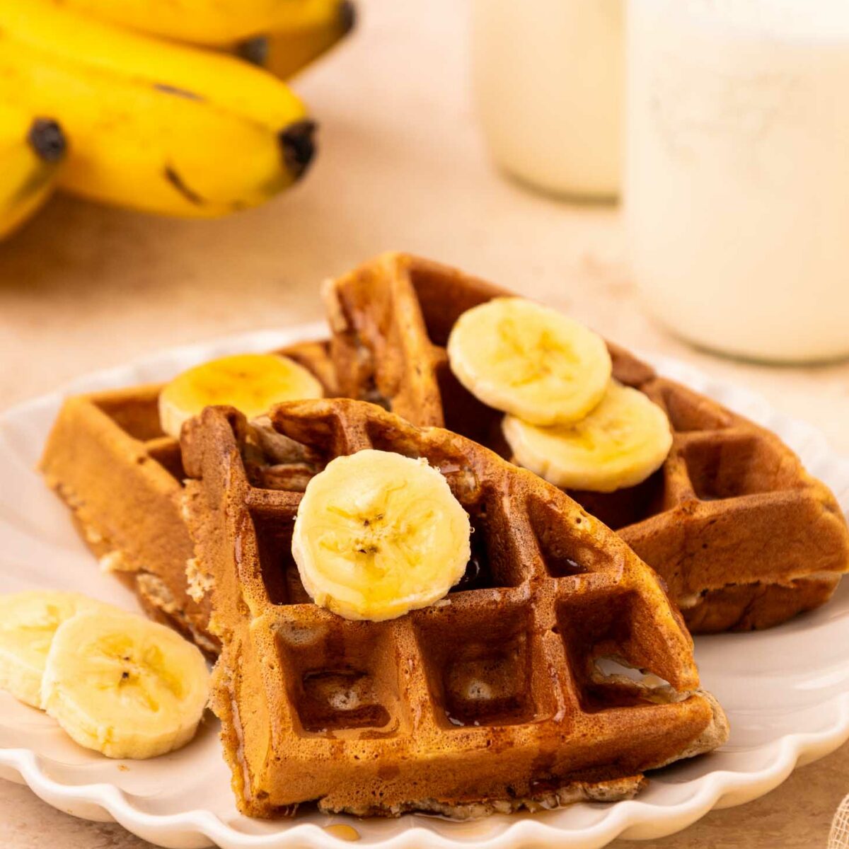 A plate with two golden brown waffles topped with banana slices, set beside a bunch of bananas and a bottle of milk. The scene suggests a cozy breakfast setting.