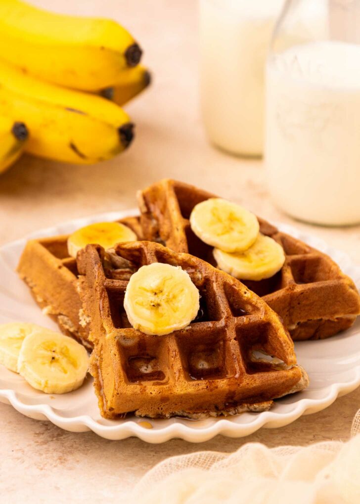 A plate with two golden brown waffles topped with banana slices, set beside a bunch of bananas and a bottle of milk. The scene suggests a cozy breakfast setting.