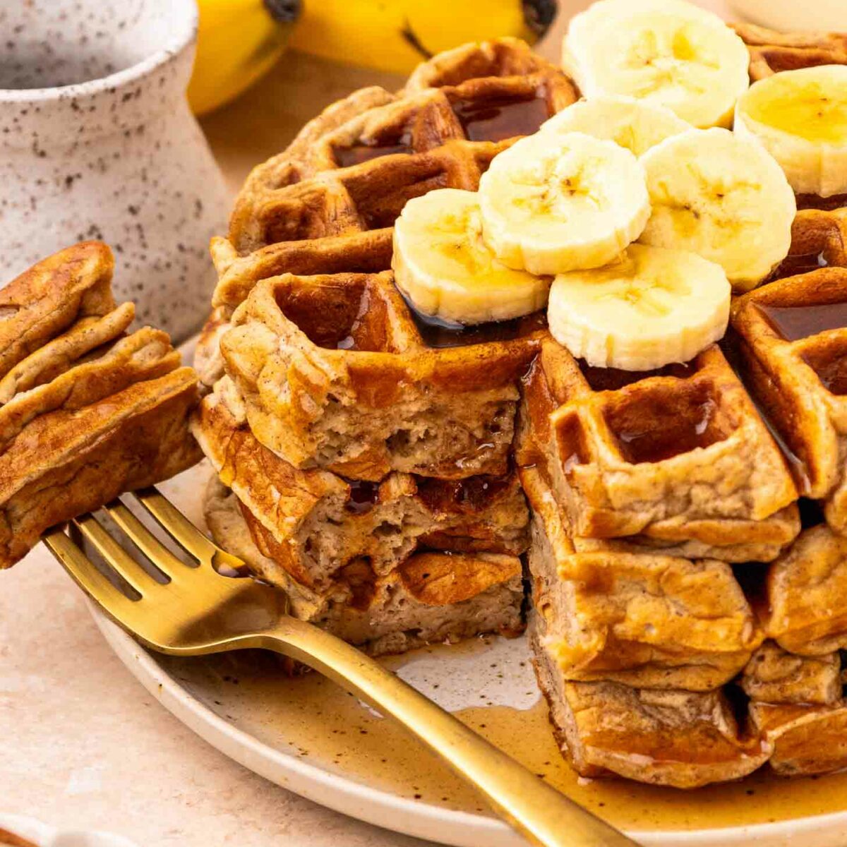 A stack of waffles topped with banana slices, with a golden fork beside them. A bunch of bananas and a ceramic cup are in the background. Syrup is drizzled over the waffles.