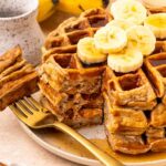 A stack of waffles topped with banana slices, with a golden fork beside them. A bunch of bananas and a ceramic cup are in the background. Syrup is drizzled over the waffles.