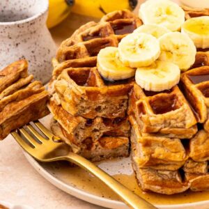 A stack of waffles topped with banana slices, with a golden fork beside them. A bunch of bananas and a ceramic cup are in the background. Syrup is drizzled over the waffles.