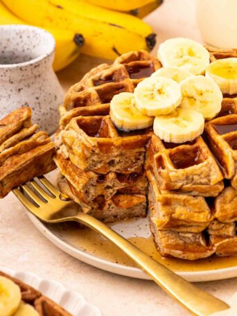 A stack of waffles topped with banana slices, with a golden fork beside them. A bunch of bananas and a ceramic cup are in the background. Syrup is drizzled over the waffles.
