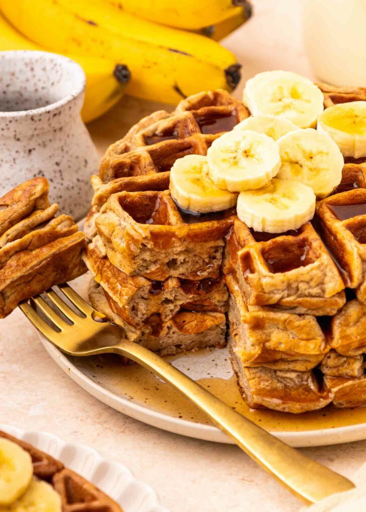 A stack of waffles topped with banana slices, with a golden fork beside them. A bunch of bananas and a ceramic cup are in the background. Syrup is drizzled over the waffles.