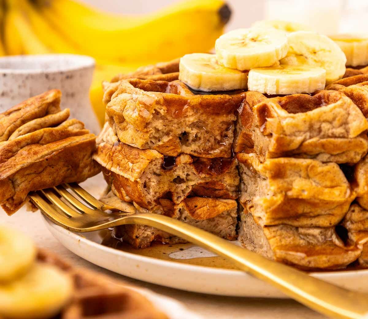 A stack of thick waffles topped with banana slices sits on a plate with a gold fork. A bite has been cut out, revealing the fluffy texture. Bananas and a small jar are blurred in the background.