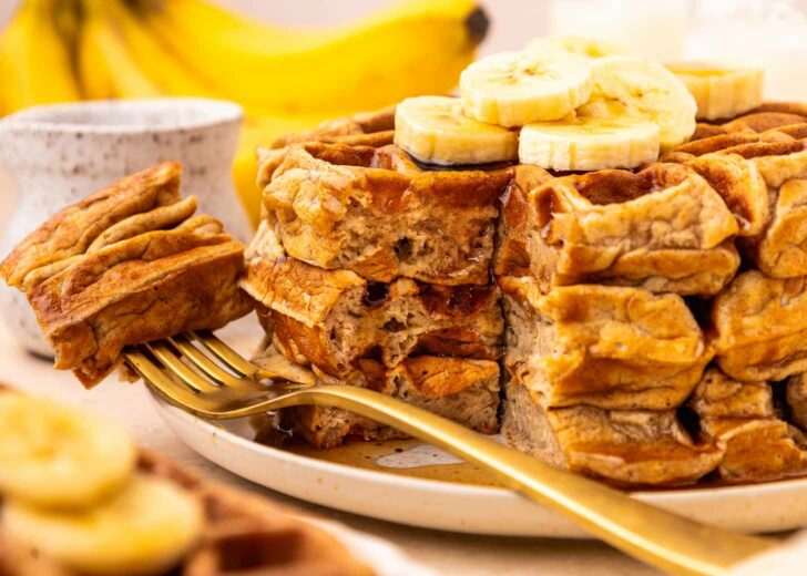 A stack of thick waffles topped with banana slices sits on a plate with a gold fork. A bite has been cut out, revealing the fluffy texture. Bananas and a small jar are blurred in the background.