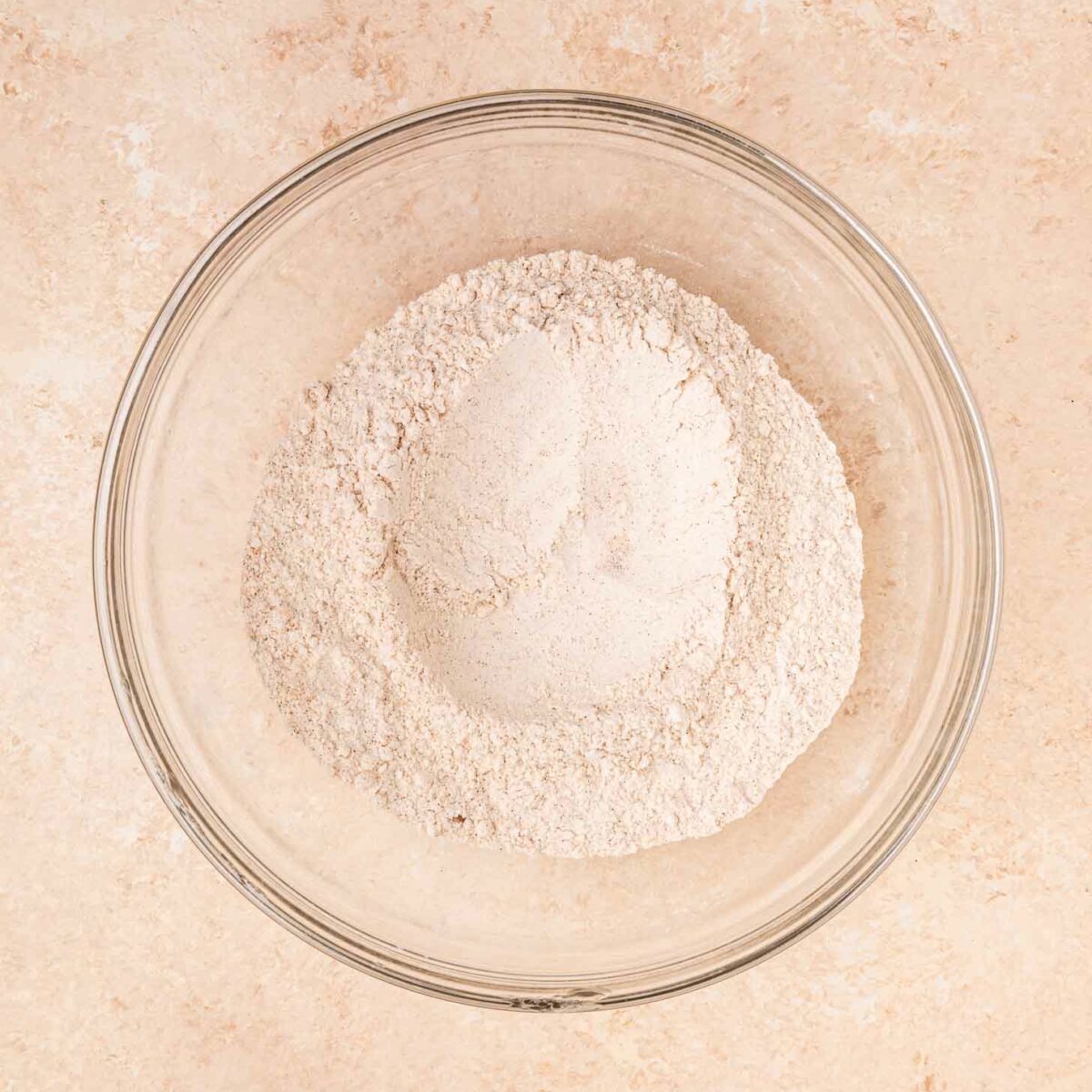 A clear glass bowl filled with flour and other dry ingredients sits on a light beige countertop.