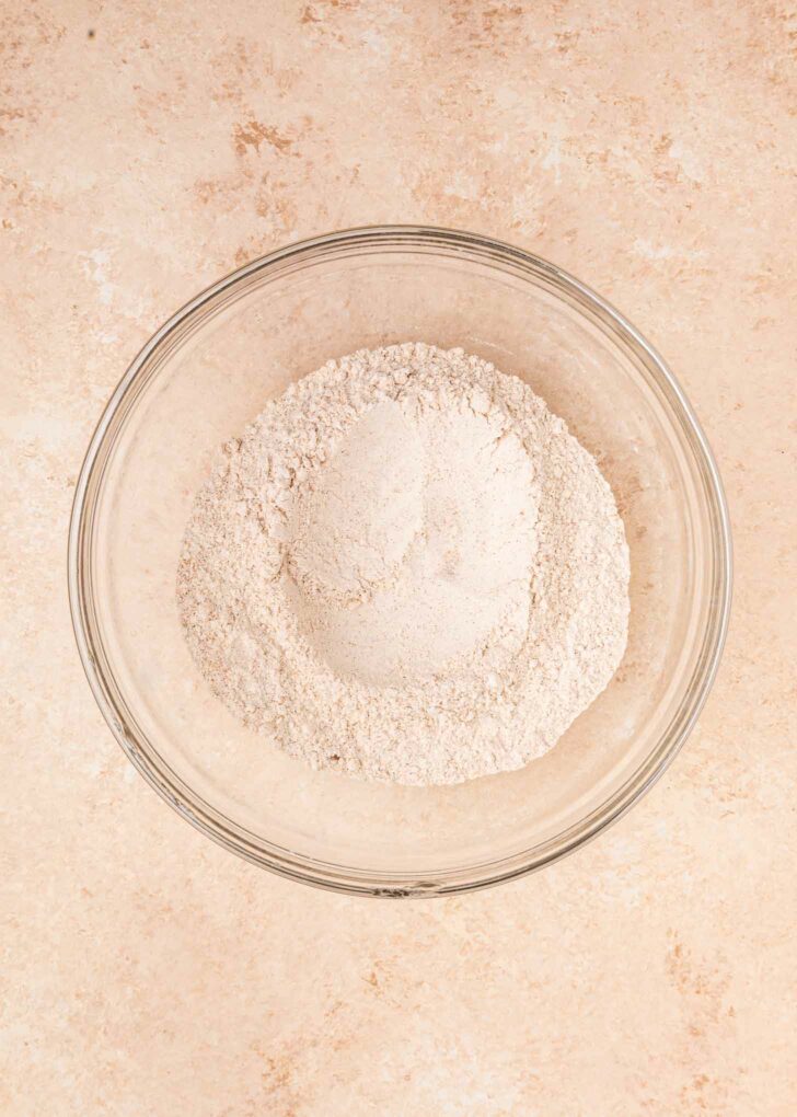 A clear glass bowl filled with flour and other dry ingredients sits on a light beige countertop.