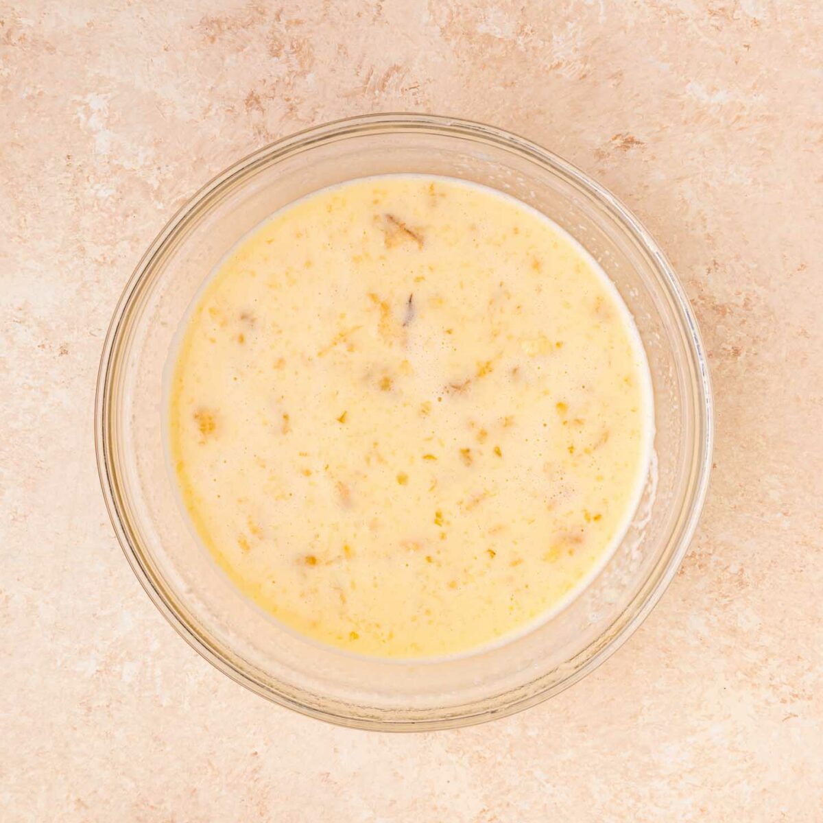 A glass bowl filled with a creamy, speckled egg mixture sits on a light beige countertop viewed from above.