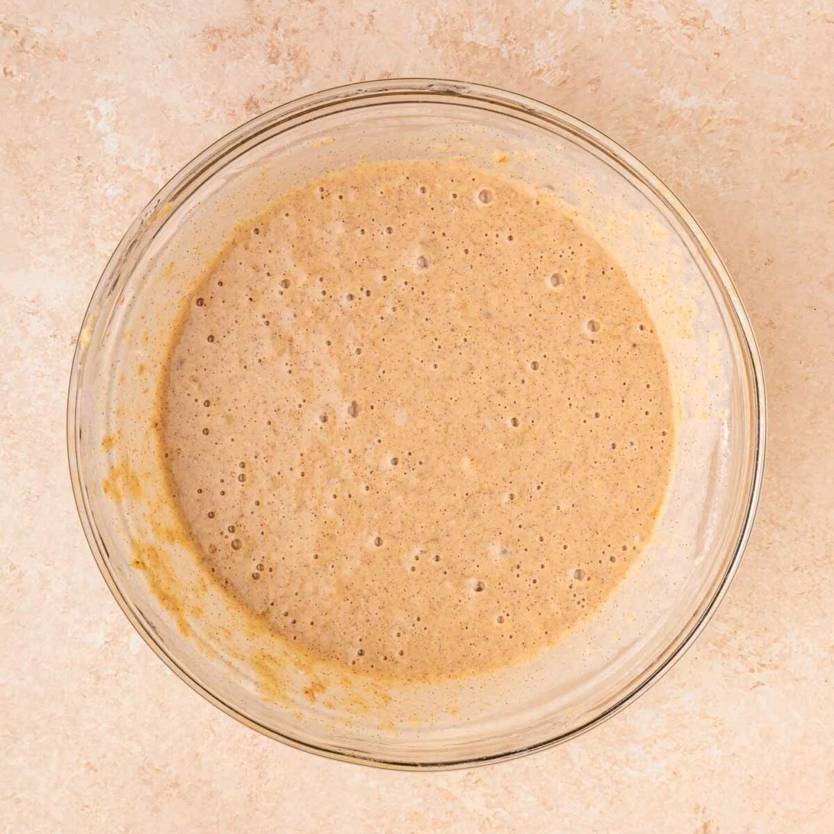 A glass bowl filled with light brown, bubbly waffle batter sits on a beige countertop.