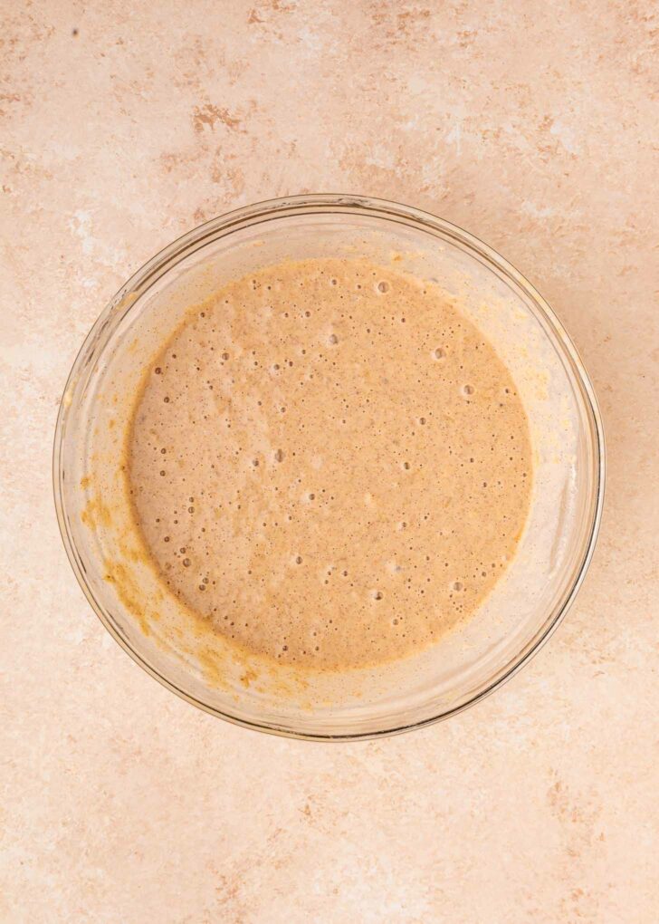 A glass bowl filled with light brown, bubbly waffle batter sits on a beige countertop.
