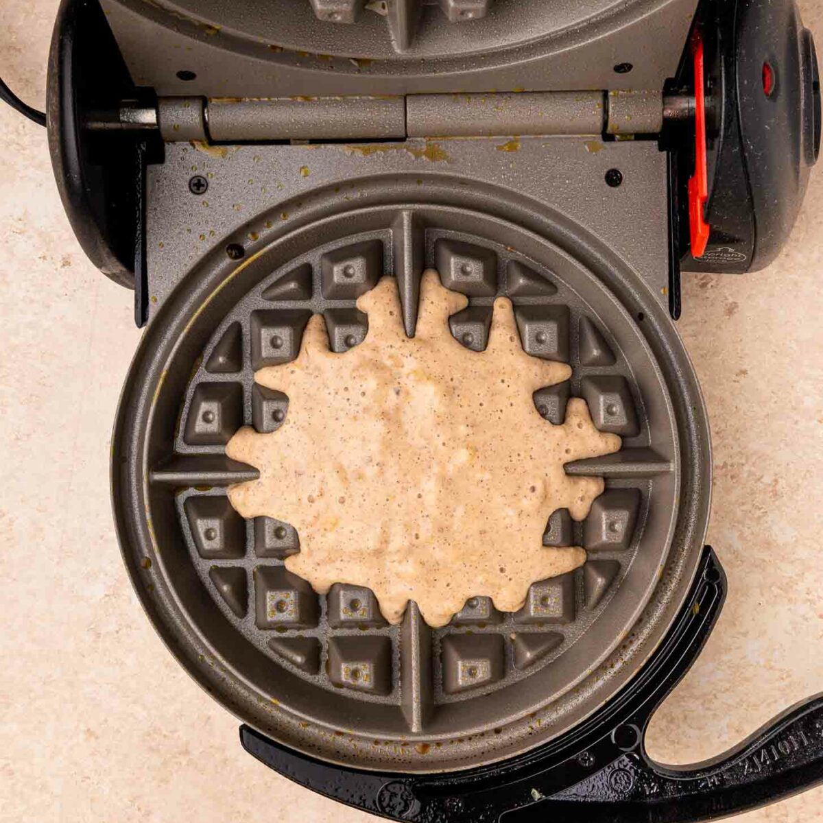 A waffle maker with its lid open shows raw waffle batter spread unevenly over the lower grid, ready to be cooked. The appliance sits on a light-colored countertop.