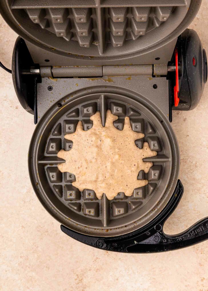 A waffle maker with its lid open shows raw waffle batter spread unevenly over the lower grid, ready to be cooked. The appliance sits on a light-colored countertop.