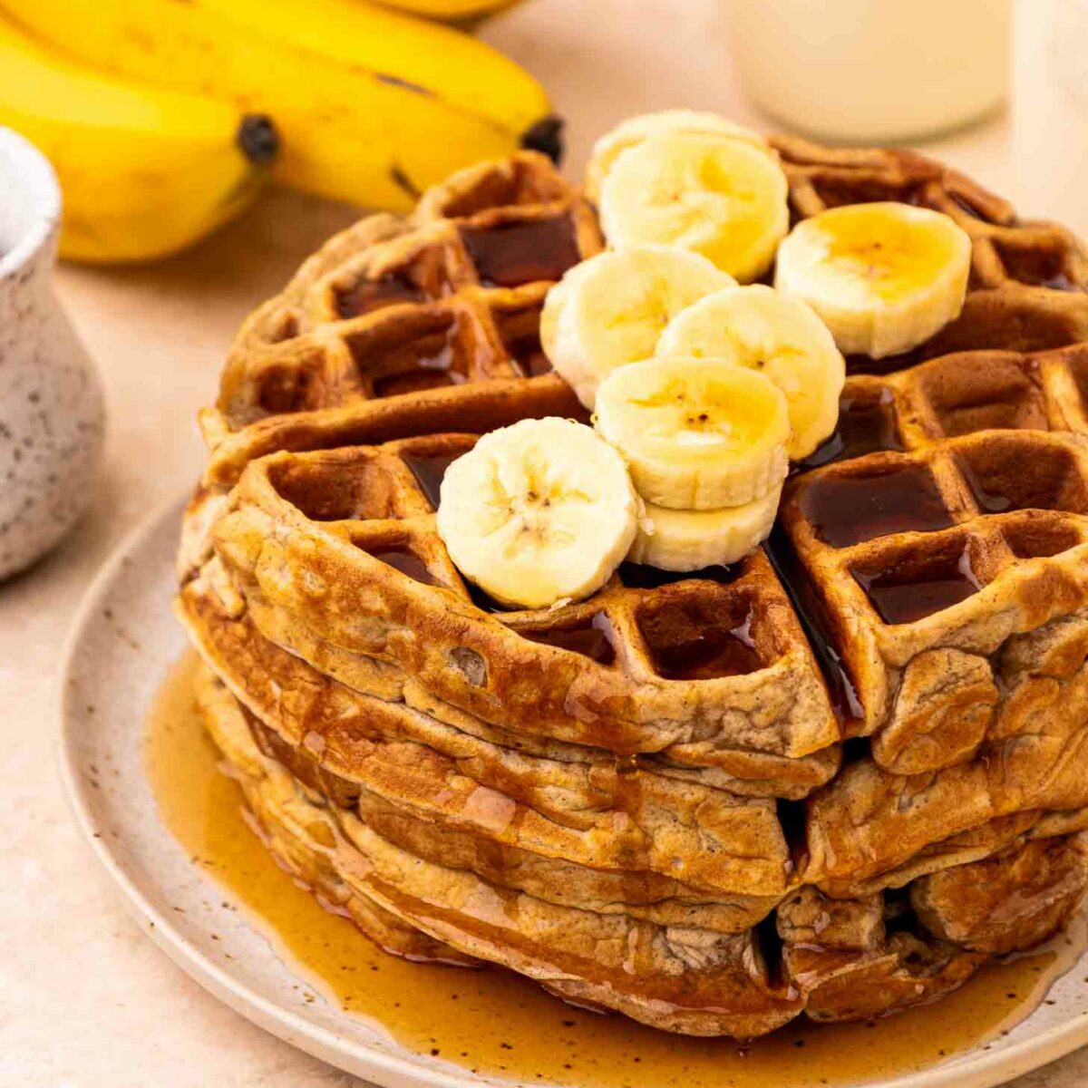 A stack of waffles topped with banana slices and syrup sits on a plate. In the background, there are whole bananas, a cup with a beverage, and a small pitcher.