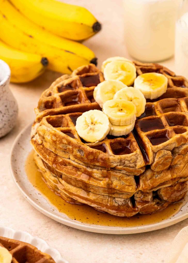 A stack of waffles topped with banana slices and syrup sits on a plate. In the background, there are whole bananas, a cup with a beverage, and a small pitcher.