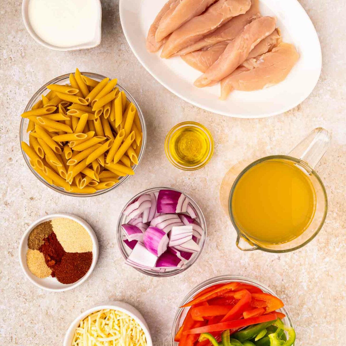 Overhead view of ingredients on a light surface: raw chicken breasts, penne pasta, sliced red and green bell peppers, chopped red onion, shredded cheese, chicken broth, spices, olive oil, and cream.