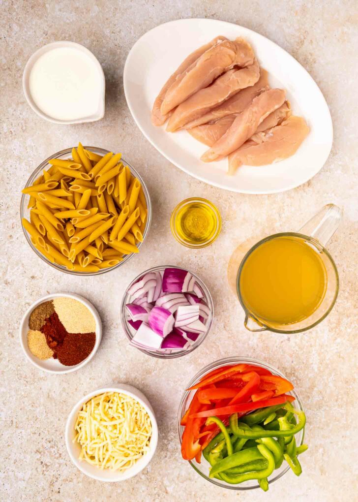 Overhead view of ingredients on a light surface: raw chicken breasts, penne pasta, sliced red and green bell peppers, chopped red onion, shredded cheese, chicken broth, spices, olive oil, and cream.