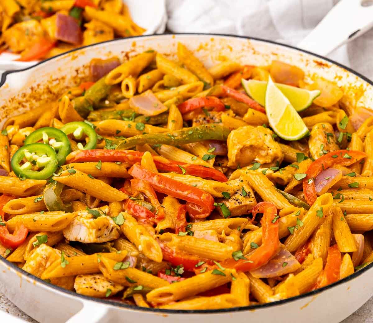 A skillet filled with penne pasta, mixed with colorful bell peppers, red onion, chunks of chicken, sliced jalapeños, and garnished with lime wedges and herbs.