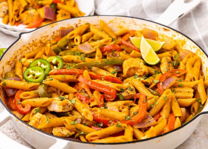 A skillet filled with penne pasta, mixed with colorful bell peppers, red onion, chunks of chicken, sliced jalapeños, and garnished with lime wedges and herbs.