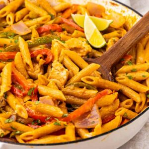 A skillet filled with creamy penne pasta, sliced red bell peppers, onions, and chicken strips, garnished with fresh herbs and lime wedges on top, with a wooden spoon resting in the pasta.
