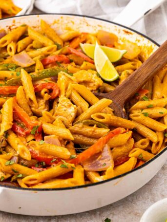 A skillet filled with creamy penne pasta, sliced red bell peppers, onions, and chicken strips, garnished with fresh herbs and lime wedges on top, with a wooden spoon resting in the pasta.