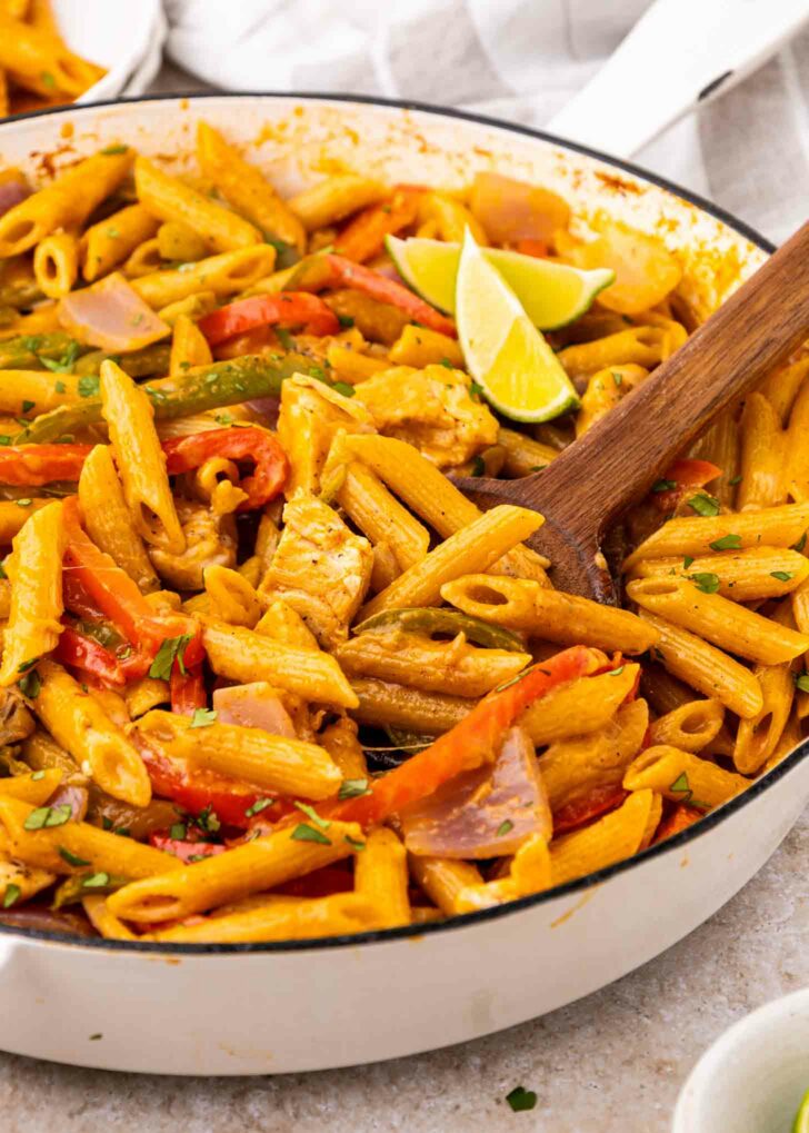 A skillet filled with creamy penne pasta, sliced red bell peppers, onions, and chicken strips, garnished with fresh herbs and lime wedges on top, with a wooden spoon resting in the pasta.