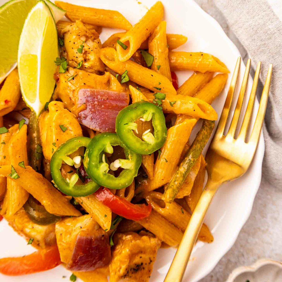 A white plate of creamy penne pasta with chicken, red onions, bell peppers, and jalapeno slices, garnished with lime wedges. A gold fork rests on the plate; some chopped herbs are nearby.