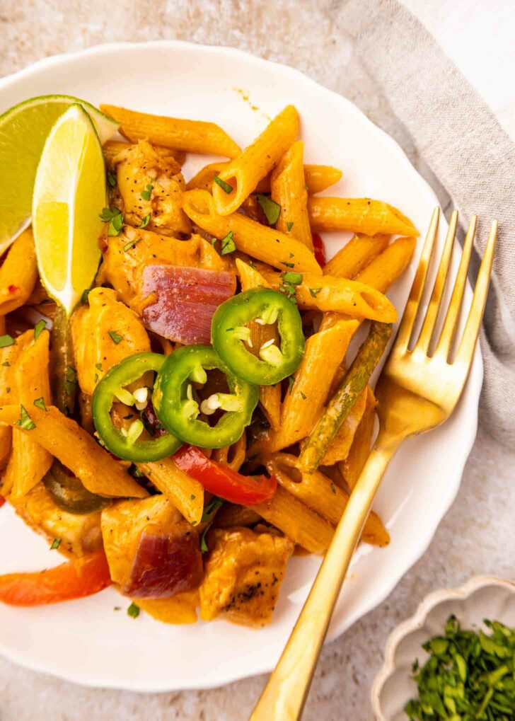 A white plate of creamy penne pasta with chicken, red onions, bell peppers, and jalapeno slices, garnished with lime wedges. A gold fork rests on the plate; some chopped herbs are nearby.