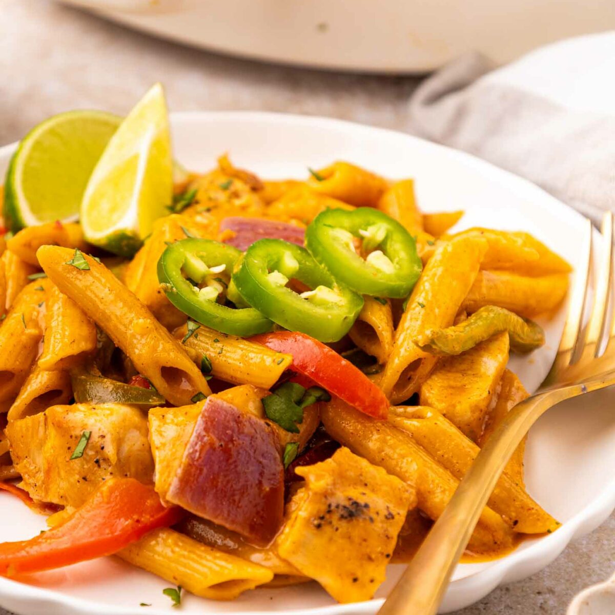 A plate of creamy pasta with penne, red onions, bell peppers, and chunks of chicken, garnished with sliced jalapeños and a lime wedge. A fork and knife rest on the plates edge.