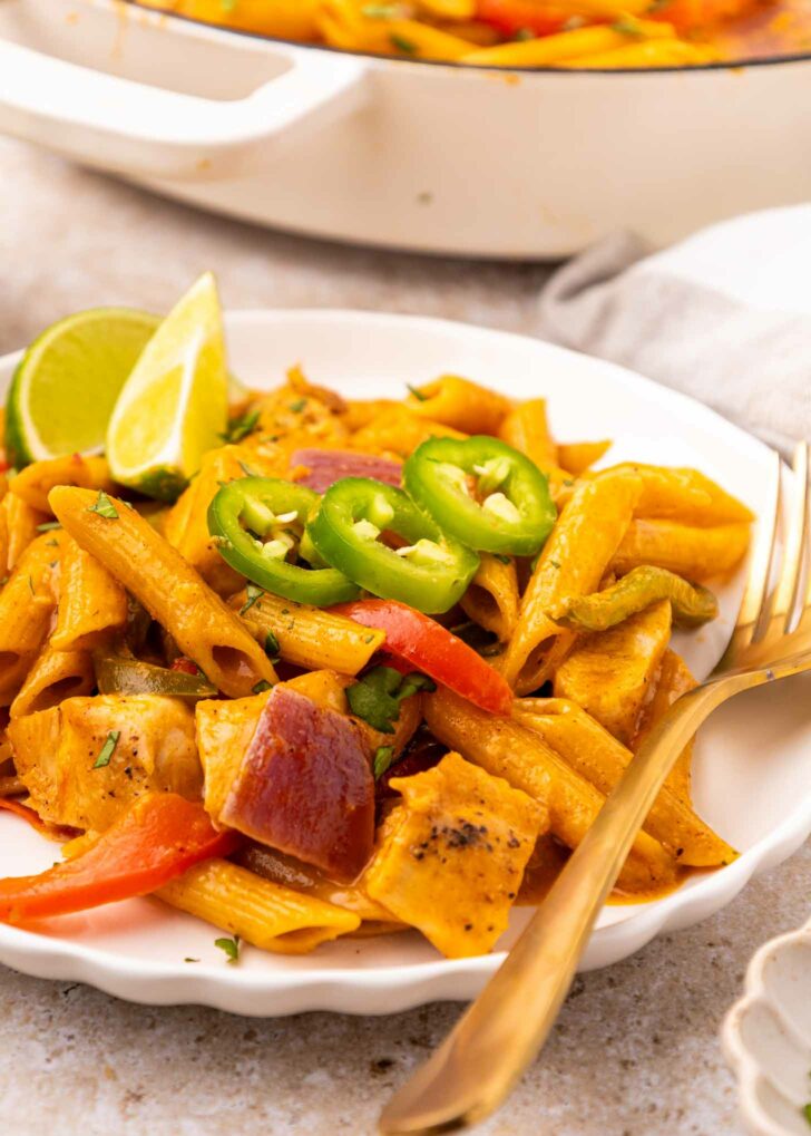 A plate of creamy pasta with penne, red onions, bell peppers, and chunks of chicken, garnished with sliced jalapeños and a lime wedge. A fork and knife rest on the plates edge.
